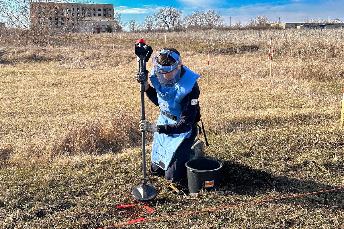 FSD deminer in blue vest, kneeling in a Kharkiv field, searches for mines during a second mine clearance survey.