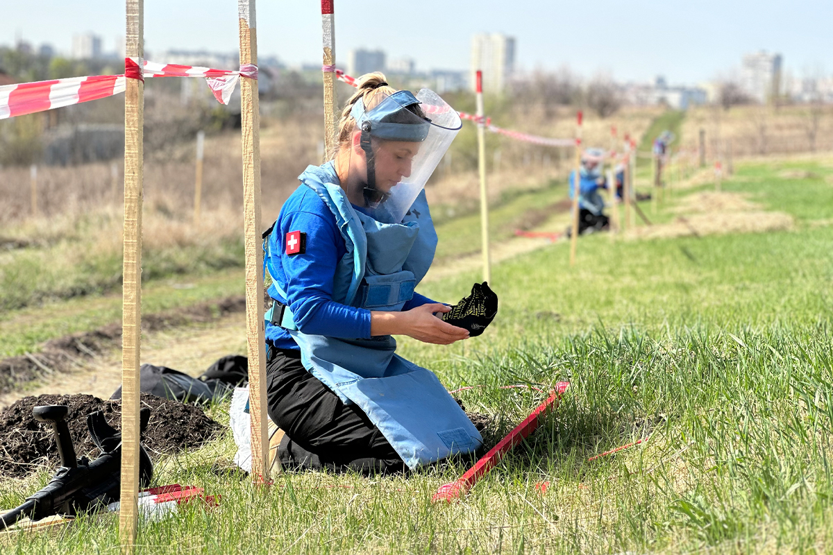 FSD female deminer in blue vest, kneeling on grass in Kharkiv field, searching for mines during second clearance training survey FSD female deminer in blue vest, kneeling on grass in Kharkiv field, searching for mines during second clearance training survey.